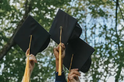 Hands raising black graduation caps with gold tassels against green trees