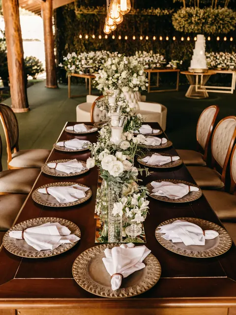 A long wooden table with gold chargers, white napkins and a runner of white flowers under hanging bulbs