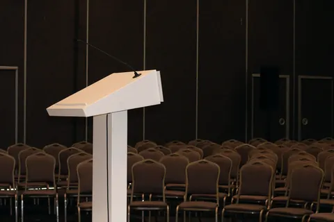 A white lectern before rows of chairs in a softly lit conference hall