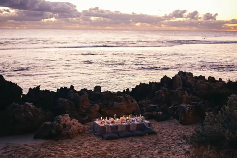 A candlelit dinner table among the rocks of the shore at sunset