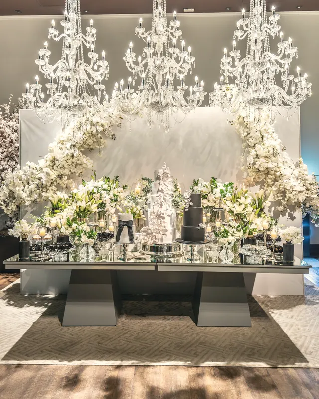 Three crystal chandeliers above a wedding dessert table drowned in white orchids and lilies