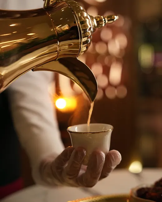A gold dallah pouring Saudi coffee into a cup held by a gloved hand