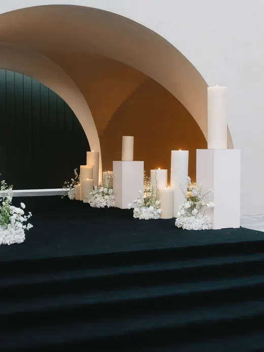 An arched entrance with tall white candles on plinths and white flowers above dark steps