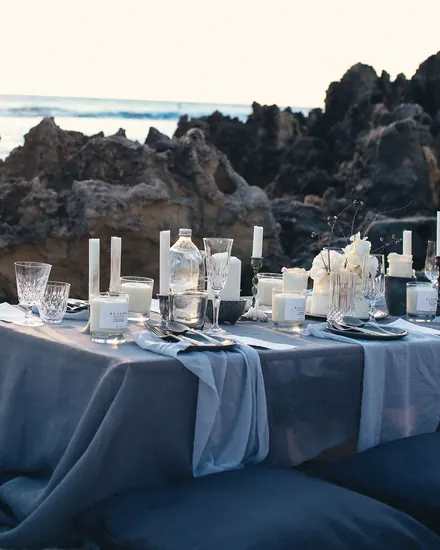 A dinner table with blue linens and white candles among the rocks of the shore