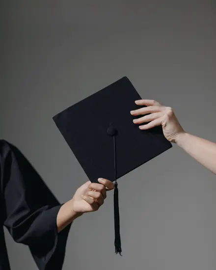 Two hands passing a black graduation cap with its tassel against a grey backdrop
