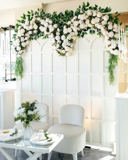 A milka corner with white panels, an arch of white roses and greenery, and two soft chairs