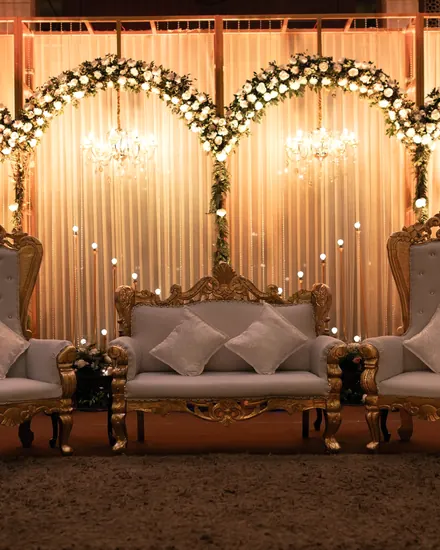 A kosha of white-flower arches, small chandeliers and a gilded sofa in warm light